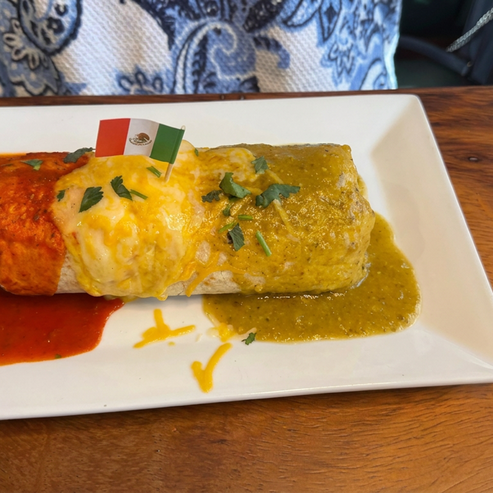 Cheesy burrito topped with cilantro and a Mexican flag, served with red and green sauces on a white plate.